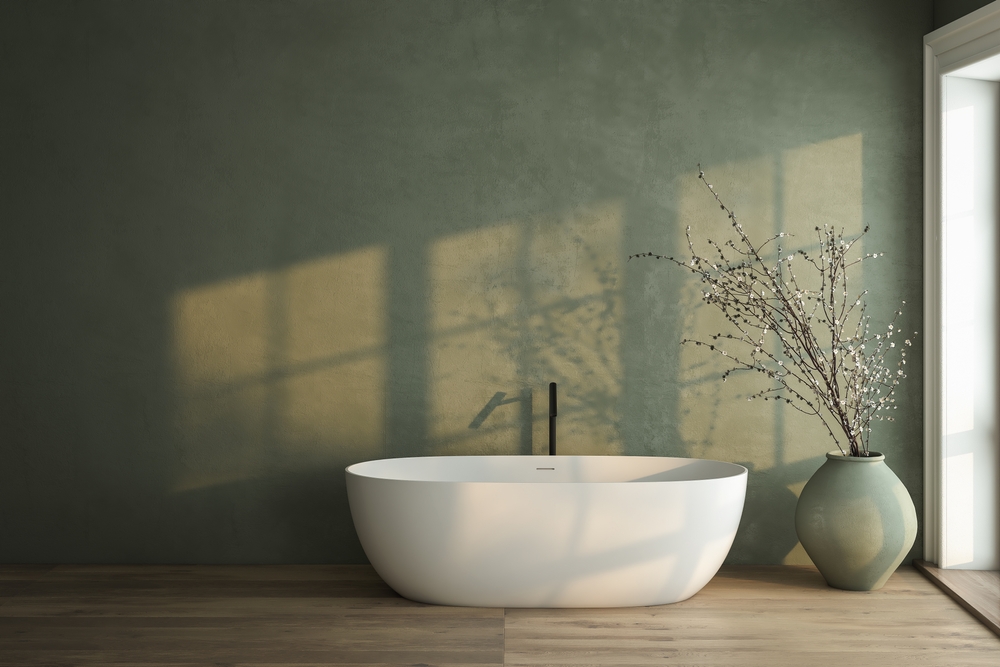 White bathtub against a textured, dark green wall with sunlit window shadows, next to a large vase with dry branches on a wood floor.