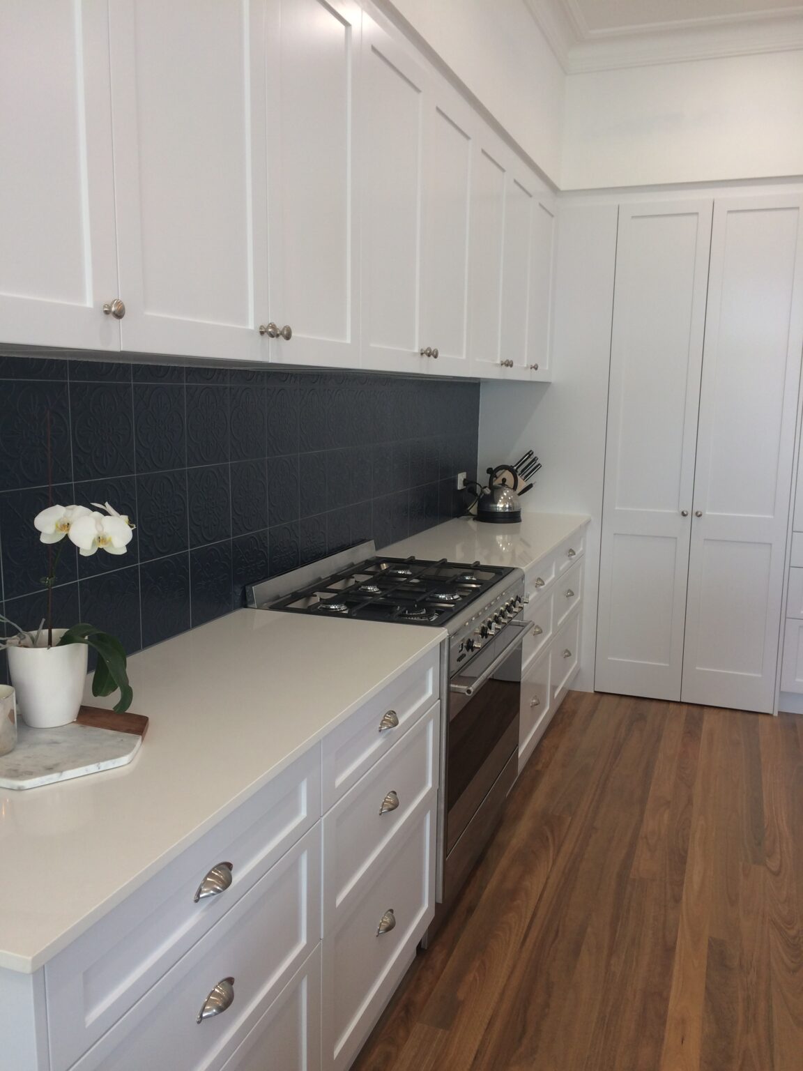 A kitchen with white shaker cabinets, a stainless steel stove, and a dark blue embossed tile backsplash on top of wood floors.